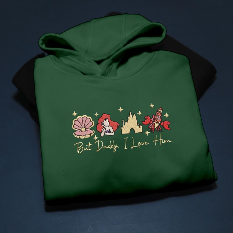 Pode incluir: Um moletom com capuz verde escuro com imagens bordadas de uma concha, uma sereia, um castelo e um caranguejo. O texto "But Daddy, I Love Him" &eacute; bordado em dourado abaixo.