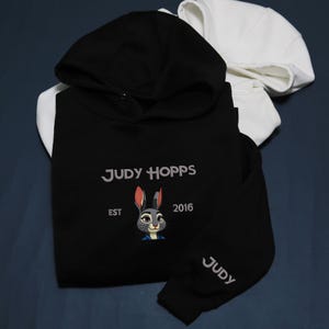 May include: Black hoodie featuring the text "JUDY HOPPS" and a cartoon rabbit face. The hoodie has a black hood and sleeves, with the word "JUDY" on the sleeve. A white hoodie is visible in the background.