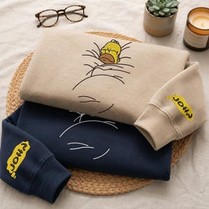 May include: A beige sweatshirt featuring a graphic of Homer Simpson sleeping under a blanket, with the text "D'OH!" embroidered in yellow on the sleeve. A second navy blue sweatshirt is layered underneath, also featuring the same yellow "D'OH!" text.
