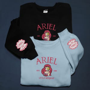 May include: Black and pale blue sweatshirts with pink text and a mermaid graphic. The black sweatshirt has a "Linda" seashell design on the sleeve. The light blue sweatshirt has the words "Little Mermaid" below the graphic. The sweatshirts are likely made of cotton.