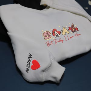 Pode incluir: Uma caneca de cer&acirc;mica branca com uma al&ccedil;a preta e o texto "But First, Coffee" em estilo manuscrito. A caneca est&aacute; sobre uma superf&iacute;cie branca. A caneca tem aproximadamente 10 cm de altura.