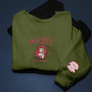May include: An olive green sweatshirt with the name "ARIEL" embroidered in pink, along with a Little Mermaid graphic. The sleeve displays a pink seashell with the name "LINDA". A black sweatshirt is visible behind it.