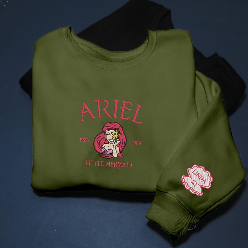 May include: An olive green sweatshirt with the name "ARIEL" embroidered in pink, along with a Little Mermaid graphic. The sleeve displays a pink seashell with the name "LINDA". A black sweatshirt is visible behind it.