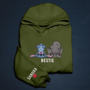 K&ouml;nnte beinhalten: Olivgr&uuml;ner Kapuzenpullover mit Kapuze und langen &Auml;rmeln. Auf der Vorderseite befindet sich ein blaues und graues Cartoon-Design mit dem Wort "BESTIE" in Wei&szlig;. Der linke &Auml;rmel tr&auml;gt den Namen "SANDRA" und ein rotes Herz.