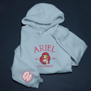May include: A pale blue hoodie with the name "ARIEL" in pink, and an image of Ariel from The Little Mermaid. The hoodie also has the text "EST. 1989" and a small embroidered design on the sleeve.
