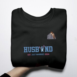 Puede incluir: Sudadera negra con la palabra "HUSBAND" en azul, con un personaje de dibujos animados y el texto "EST. JUST MARRIED 2024". Una peque&ntilde;a ilustraci&oacute;n de una pareja est&aacute; en la parte superior derecha. La sudadera est&aacute; doblada.