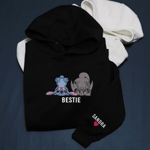 K&ouml;nnte beinhalten: Schwarzer Kapuzenpullover mit Kapuze, bestickt mit einem Design von zwei Zeichentrickfiguren und dem Wort "BESTIE" in Wei&szlig;. Der &Auml;rmel hat den Namen "SANDRA" und ein rotes Herz. Im Hintergrund ist ein wei&szlig;er Kapuzenpullover zu sehen.