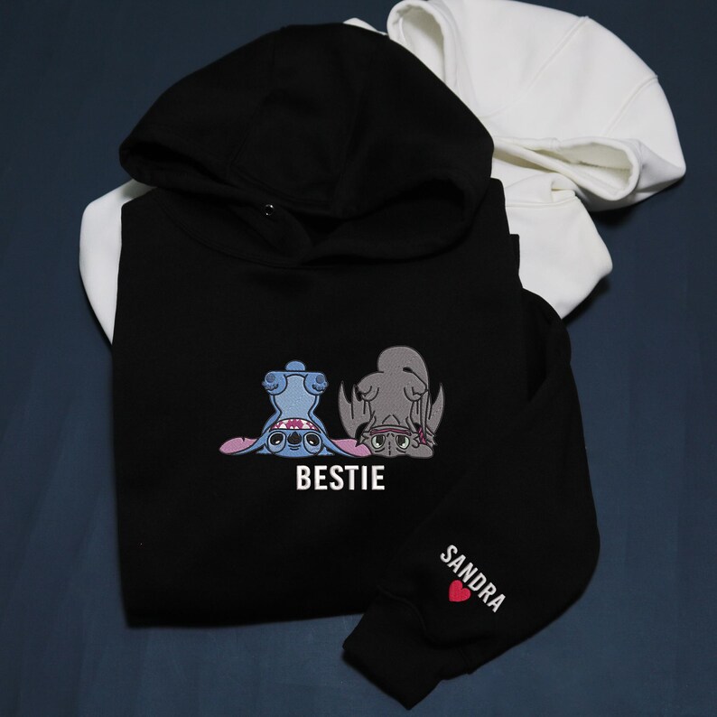 K&ouml;nnte beinhalten: Schwarzer Kapuzenpullover mit Kapuze, bestickt mit einem Design von zwei Zeichentrickfiguren und dem Wort "BESTIE" in Wei&szlig;. Der &Auml;rmel hat den Namen "SANDRA" und ein rotes Herz. Im Hintergrund ist ein wei&szlig;er Kapuzenpullover zu sehen.