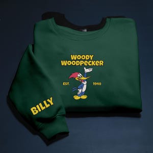 May include: A dark green sweatshirt featuring the cartoon character Woody Woodpecker. The design includes the text "WOODY WOODPECKER" in yellow, the year "1940", and the word "BILLY" on the sleeve.