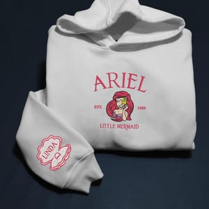 May include: White hooded sweatshirt with long sleeves. The front displays the word "ARIEL" in pink script above a mermaid illustration. Below the illustration are the words "EST. 1989 LITTLE MERMAID". The left sleeve has a pink embroidered name.