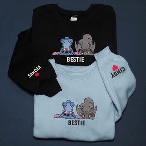 K&ouml;nnte beinhalten: Schwarze und hellblaue Sweatshirts mit einem Cartoon-Charakter-Design und dem Wort "BESTIE" darunter. Ein &Auml;rmel hat den Namen "SANDRA" und ein Herz, der andere "CINDY" und ein Herz. Ein passendes Set.