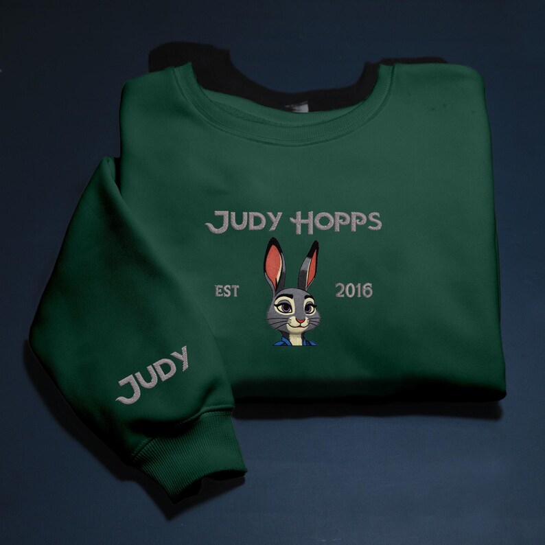 May include: A dark green sweatshirt featuring the embroidered text "JUDY HOPPS" in white, with a cartoon rabbit image beneath. The sleeve displays "JUDY" in white embroidery. The year "2016" is also embroidered on the sweatshirt.