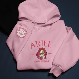 May include: A pale pink hoodie with the name "ARIEL" embroidered in a script font, and an illustration of Ariel. The text "LITTLE MERMAID" is also visible. The sleeve has a patch that says "LINDA".