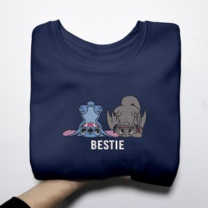 K&ouml;nnte beinhalten: Marineblaues Sweatshirt mit einem Cartoon-Design von zwei Charakteren, einem blauen und einem grauen, mit dem Wort "BESTIE" in Wei&szlig;. Die Charaktere sind auf dem Kopf. Das Sweatshirt ist gefaltet.
