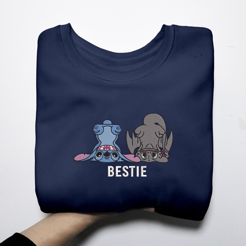 K&ouml;nnte beinhalten: Marineblaues Sweatshirt mit einem Cartoon-Design von zwei Charakteren, einem blauen und einem grauen, mit dem Wort "BESTIE" in Wei&szlig;. Die Charaktere sind auf dem Kopf. Das Sweatshirt ist gefaltet.