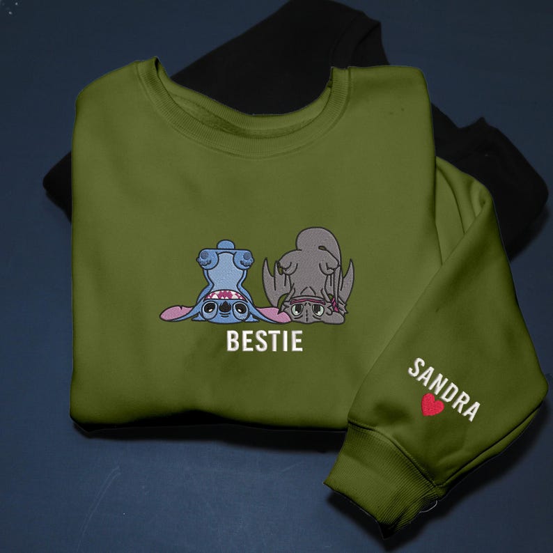 K&ouml;nnte beinhalten: Ein olivgr&uuml;nes Sweatshirt mit einer Cartoon-Grafik von zwei Figuren und dem Wort "BESTIE" in Wei&szlig;. Der &Auml;rmel zeigt den Namen "SANDRA" in Wei&szlig; mit einem roten Herz.