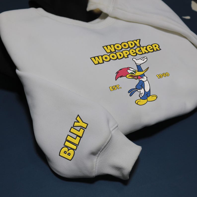 May include: White sweatshirt featuring the cartoon character Woody Woodpecker in a colorful graphic. The text "WOODY WOODPECKER" and "BILLY" are printed in yellow. The sweatshirt has a black hood and is on a dark blue surface.