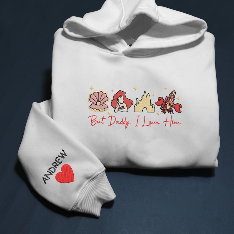 Pode incluir: Moletom branco com capuz com cord&atilde;o e punhos canelados. A frente apresenta um desenho bordado de uma concha, uma sereia, um castelo e um caranguejo, com o texto "But Daddy, I Love Him". A manga esquerda tem o nome "ANDREW" e um cora&ccedil;&atilde;o vermelho.
