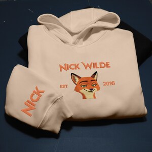 May include: Cream-coloured hoodie featuring the embroidered text "NICK WILDE" in orange, a fox graphic, and the year "2016". The sleeve displays the name "NICK" in orange. The hoodie has a hood with a drawstring.