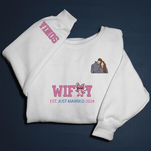 Puede incluir: Sudadera blanca con el nombre "SONIA" en rosa en la manga. La parte delantera presenta la palabra "WIFEY" en rosa, un personaje de dibujos animados y el texto "EST. JUST MARRIED 2024" en rosa y azul. Tambi&eacute;n se representa una pareja.