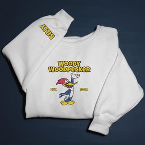 May include: White sweatshirt featuring the cartoon character Woody Woodpecker. The design includes the text "WOODY WOODPECKER" above the character, with "EST. 1940" below. The sleeve has the name "BILLY" in yellow.