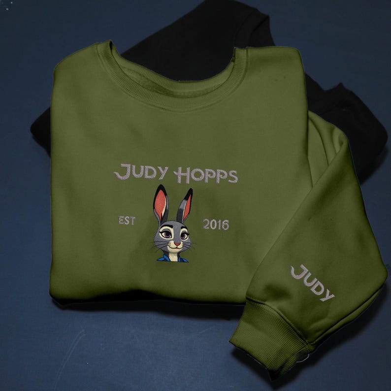 May include: An olive green sweatshirt with the name "JUDY HOPPS" embroidered in white, a bunny cartoon, and the text "EST 2016". The sleeve also has the name "JUDY" embroidered in white. A black sweatshirt is visible in the background.