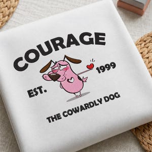 May include: A white sweatshirt featuring an embroidered graphic of a pink dog with brown ears, holding a small red heart. The text reads "COURAGE EST. 1999 THE COWARDLY DOG" in black lettering, with the character design centred on the fabric.