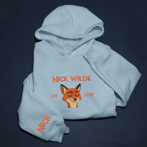May include: Pale blue hoodie featuring the embroidered text "NICK WILDE" and a cartoon fox face. The sleeve has the name "NICK" embroidered in orange. The hoodie is folded and has a hood.