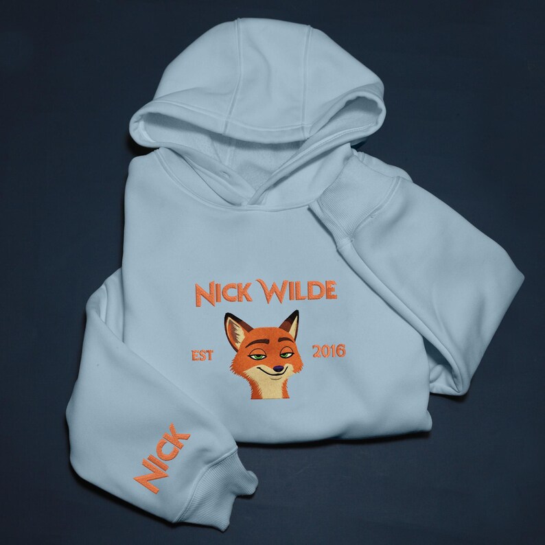 May include: Pale blue hoodie featuring the embroidered text "NICK WILDE" and a cartoon fox face. The sleeve has the name "NICK" embroidered in orange. The hoodie is folded and has a hood.