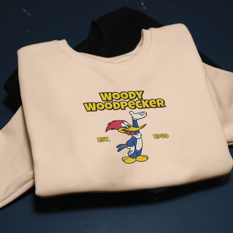 May include: A cream-colored sweatshirt featuring the cartoon character Woody Woodpecker. The design includes the text "WOODY WOODPECKER" in yellow, the year "1940", and the word "EST." The sweatshirt is layered over a black sweatshirt.