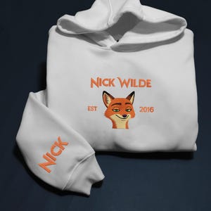 May include: White hoodie with orange embroidered text and a fox graphic. The front features "NICK WILDE" above a fox face, with "EST 2016" below. The sleeve has "NICK" embroidered in orange.