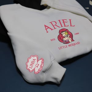 May include: Cream-coloured sweatshirt with pink embroidery. It features the name "ARIEL" above a cartoon image of Ariel from The Little Mermaid, with the text "EST. 1989 LITTLE MERMAID". The sleeve has a pink floral design with the name "LINDA".