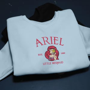 May include: A pale blue sweatshirt with the name "ARIEL" embroidered in pink, accompanied by a graphic of Ariel from The Little Mermaid. The design includes the text "EST. 1989" and "LITTLE MERMAID" in pink. A black sweatshirt is partially visible in the background.