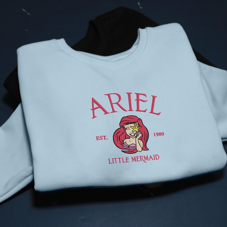 May include: A pale blue sweatshirt with the name "ARIEL" embroidered in pink, accompanied by a graphic of Ariel from The Little Mermaid. The design includes the text "EST. 1989" and "LITTLE MERMAID" in pink. A black sweatshirt is partially visible in the background.
