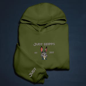 May include: An olive green hoodie with the embroidered text "JUDY HOPPS" and a cartoon rabbit design. The sleeve displays the name "JUDY". The hoodie also features the text "EST 2016".