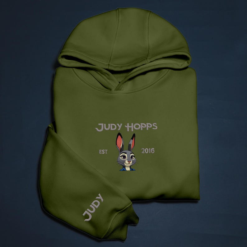 May include: An olive green hoodie with the embroidered text "JUDY HOPPS" and a cartoon rabbit design. The sleeve displays the name "JUDY". The hoodie also features the text "EST 2016".