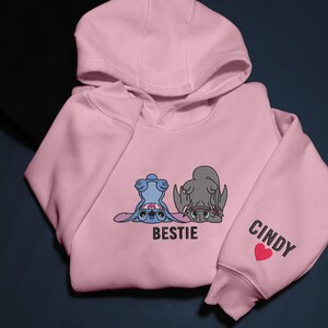 K&ouml;nnte beinhalten: Ein hellrosa Kapuzenpullover mit gestickten Cartoon-Figuren Stitch und Ohnezahn, darunter das Wort "BESTIE". Der rechte &Auml;rmel zeigt den Namen "CINDY" und ein rotes Herz. Der Hoodie hat eine Kapuze und eine K&auml;ngurutasche.