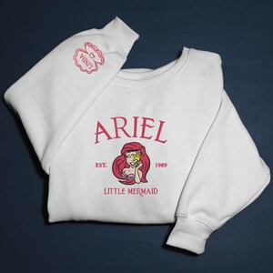 May include: White sweatshirt with a pink graphic of Ariel from The Little Mermaid, with the text "ARIEL" above the image and "EST. 1989 LITTLE MERMAID" below. The sleeve has a pink seashell design with the name "LINDA".