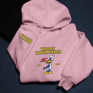 May include: A light pink hooded sweatshirt featuring the cartoon character Woody Woodpecker. The design includes the text "WOODY WOODPECKER" above the character, with "EST. 1940" below. The sleeve has the name "BILLY" in yellow.