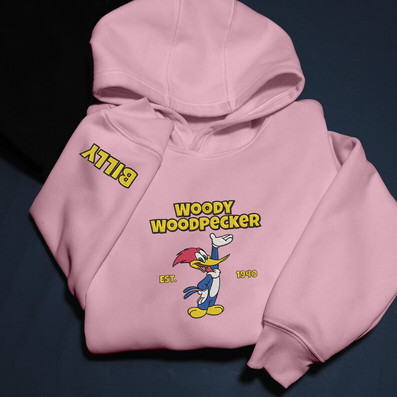 May include: A light pink hooded sweatshirt featuring the cartoon character Woody Woodpecker. The design includes the text "WOODY WOODPECKER" above the character, with "EST. 1940" below. The sleeve has the name "BILLY" in yellow.
