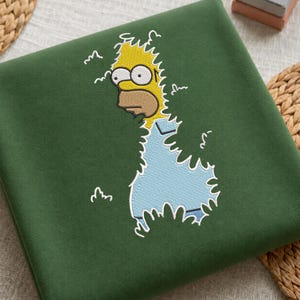 May include: A dark green sweatshirt featuring an embroidered graphic of a yellow cartoon character with wide eyes and a light blue shirt, depicted as if emerging from a dense green hedge with white outlines surrounding the character's form.