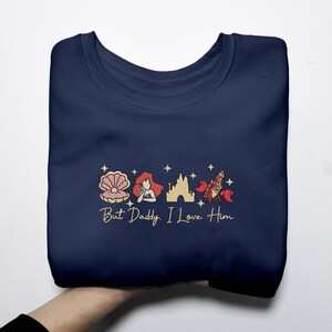 Pode incluir: Camisola azul marinho com um design bordado a ouro com uma concha, uma sereia, um castelo e um caranguejo. O texto "But Daddy, I Love Him" est&aacute; bordado por baixo. A camisola est&aacute; dobrada.