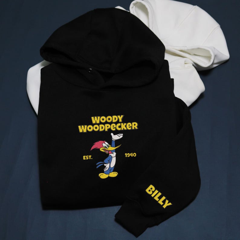 May include: Black hoodie featuring the Woody Woodpecker cartoon character with the text "WOODY WOODPECKER" and "EST. 1940". The sleeve has the name "BILLY" in yellow. A white hoodie is visible in the background.