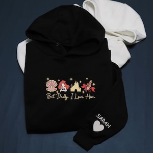 Pode incluir: Moletom preto com interior do capuz branco, apresentando um design dourado de concha, sereia, castelo e caranguejo com o texto "But Daddy, I Love Him." A manga tem o nome "SARAH" e um cora&ccedil;&atilde;o branco.