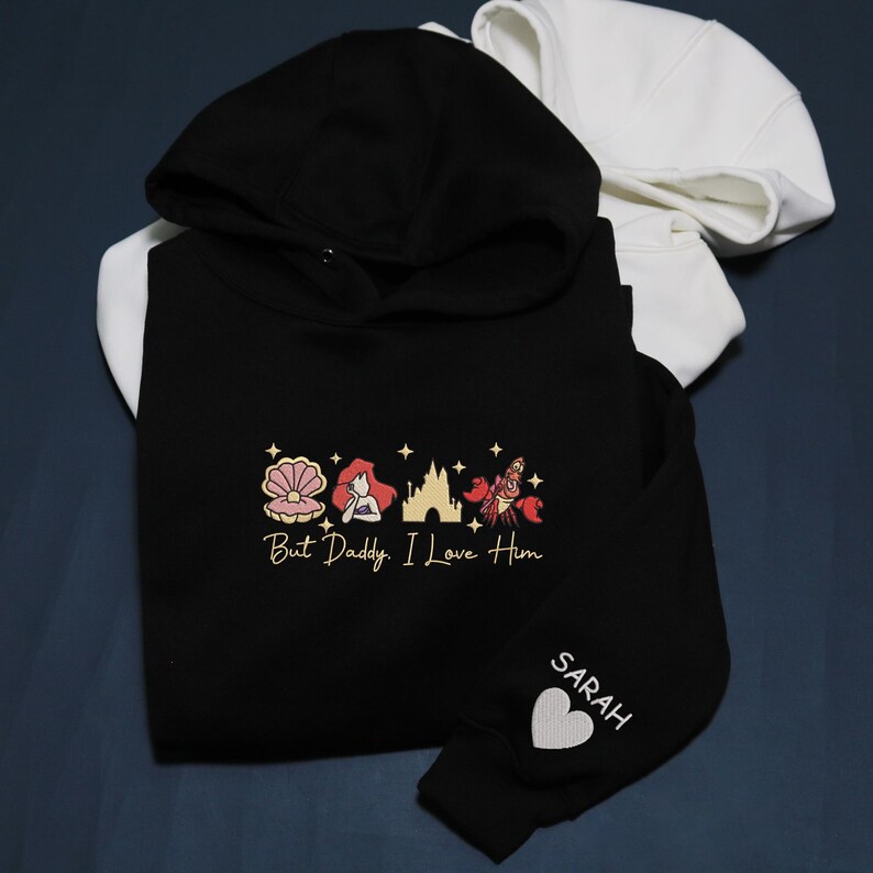 Pode incluir: Moletom preto com interior do capuz branco, apresentando um design dourado de concha, sereia, castelo e caranguejo com o texto "But Daddy, I Love Him." A manga tem o nome "SARAH" e um cora&ccedil;&atilde;o branco.