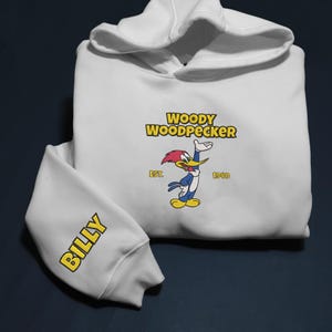 May include: White hoodie featuring the Woody Woodpecker logo in yellow and red. The sleeve displays the name "BILLY" in yellow. The hoodie has a drawstring hood and ribbed cuffs.