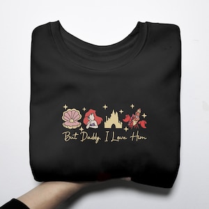 Pode incluir: Camisola preta com um design bordado com uma concha, uma sereia ruiva, um castelo e um caranguejo vermelho. O texto "But Daddy, I Love Him" est&aacute; abaixo das imagens.