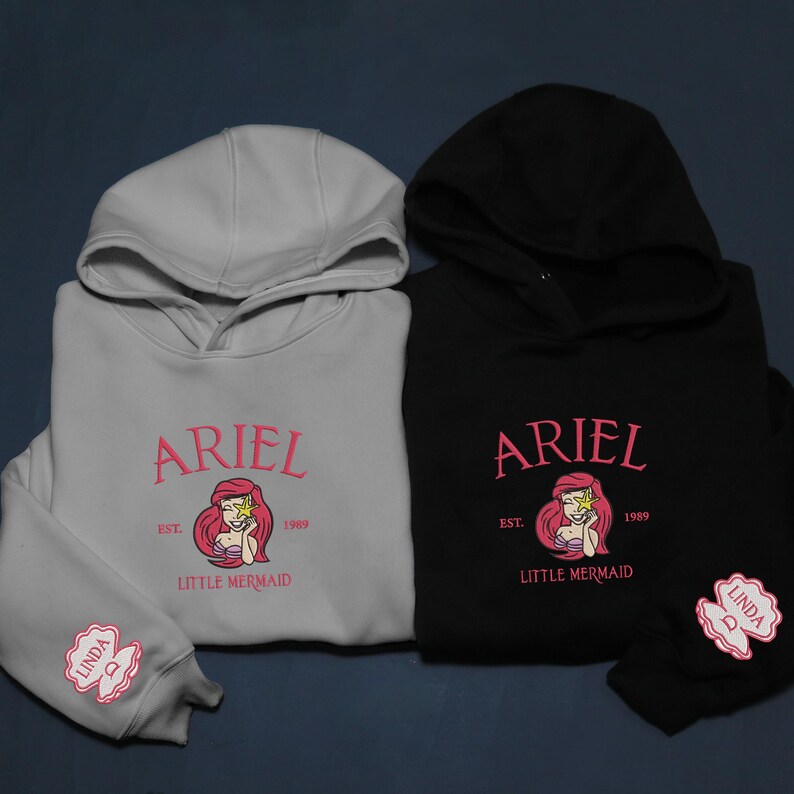 May include: Two hooded sweatshirts, one light grey and one black, with the name "ARIEL" in pink script. Below is a mermaid graphic and the text "EST. 1989 LITTLE MERMAID". The light grey sweatshirt has "LINDA" embroidered on the sleeve.