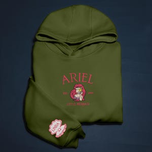 May include: An olive green hoodie with the name "ARIEL" in pink script, a graphic of Ariel, and the text "EST. 1989" and "LITTLE MERMAID". The sleeve displays a patch with the name "LINDA" in a seashell design. A casual, comfortable garment.