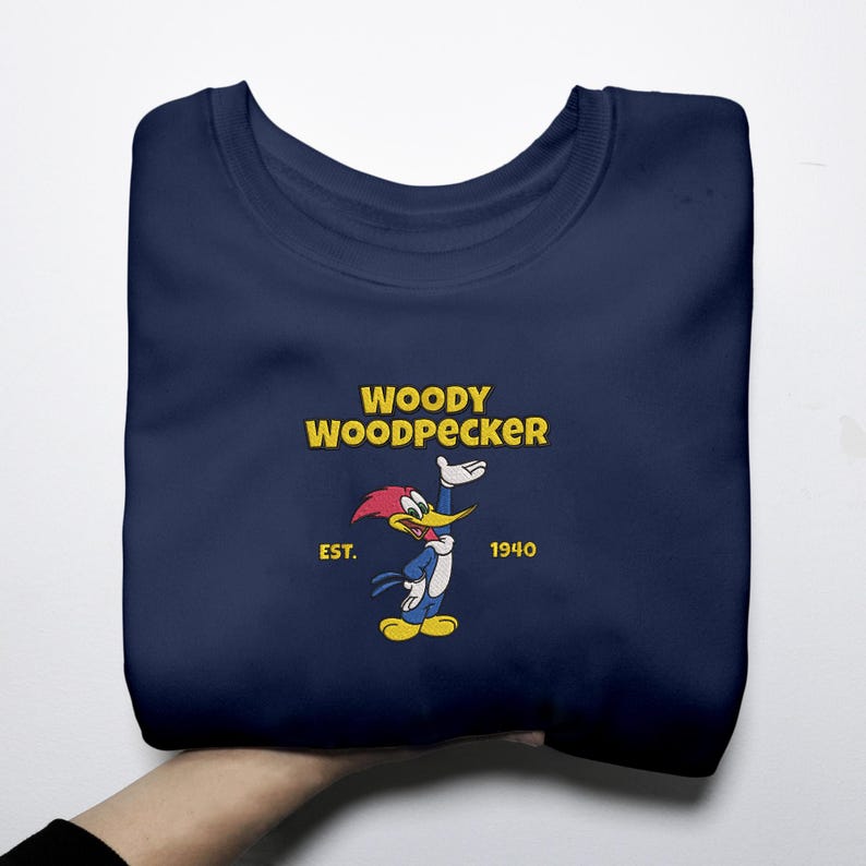 May include: Navy blue sweatshirt featuring the cartoon character Woody Woodpecker. The design includes Woody in a blue outfit with a red head and yellow beak, along with the text "WOODY WOODPECKER" and "EST. 1940" in yellow.